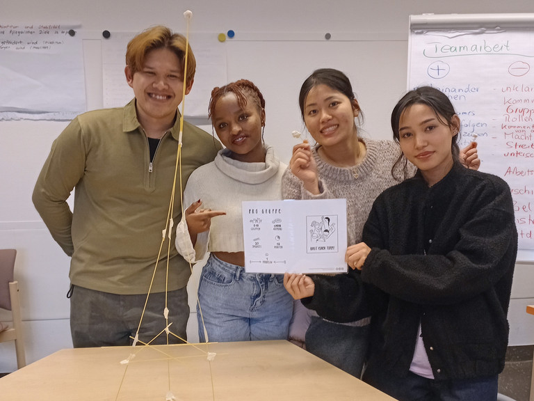 Vier Auszubildende präsentieren im Klassenraum das Ergebnis einer Teamaufgabe: einen freistehenden Turm aus Spaghetti und Marshmallows auf einem Tisch.